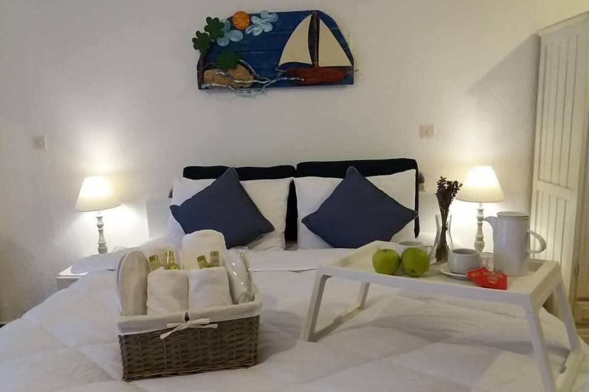 Una cama bien hecha con dos almohadas, una mesita de noche con una lámpara, una bandeja con manzanas y un objeto rojo, y un cuadro de un velero en la pared.