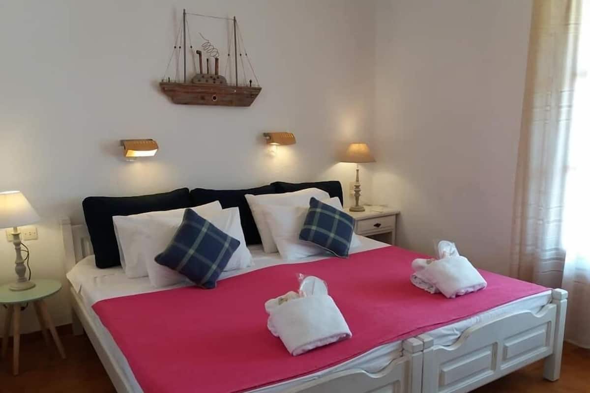 Una cama bien hecha con una colcha rosa, almohadas blancas y dos toallas. Hay dos lámparas de noche, un adorno de barco en la pared y una mesita redonda con una lámpara.