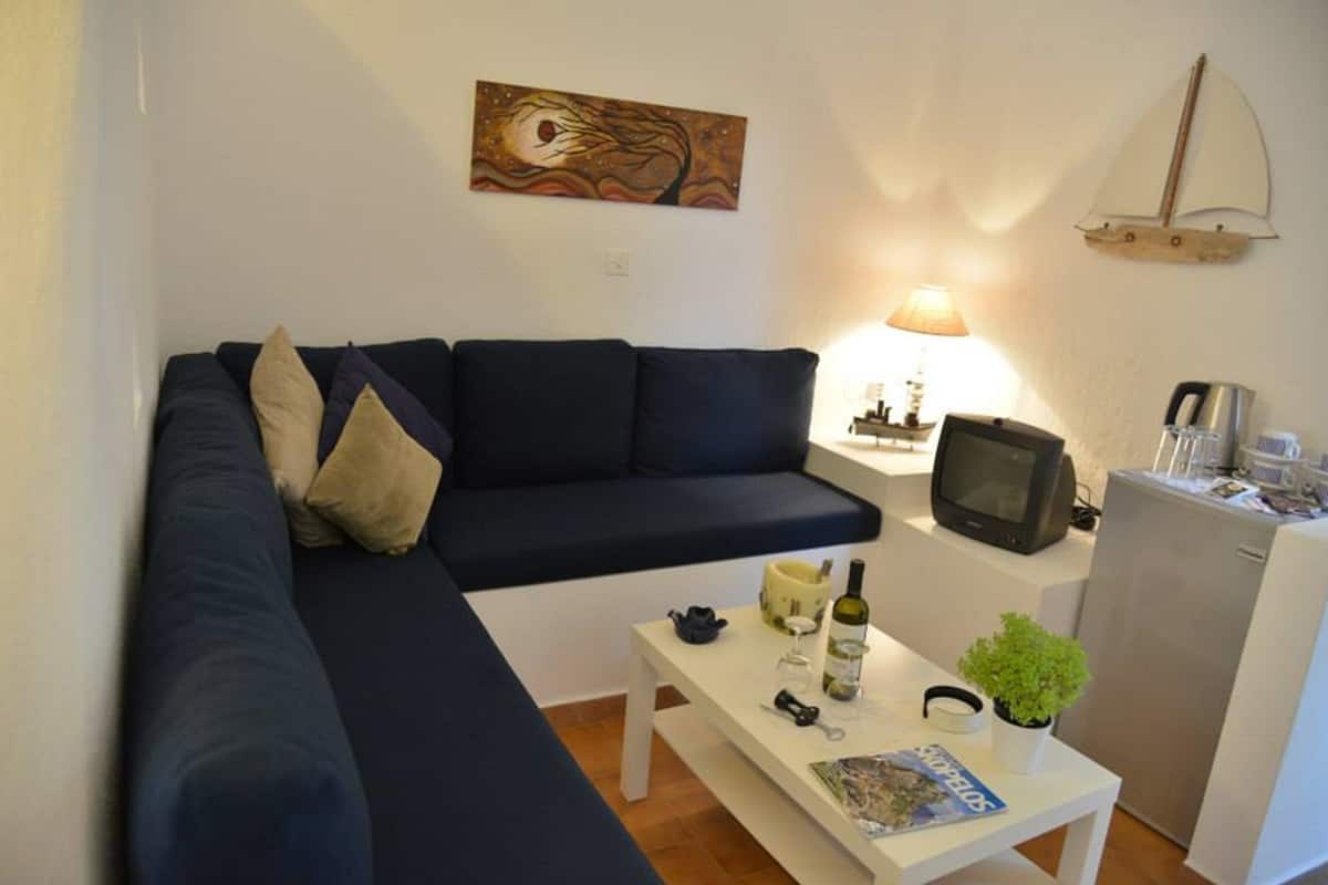 Un salón con un sofá azul oscuro, una mesa de centro blanca con una revista, una planta pequeña y un televisor sobre un mueble.