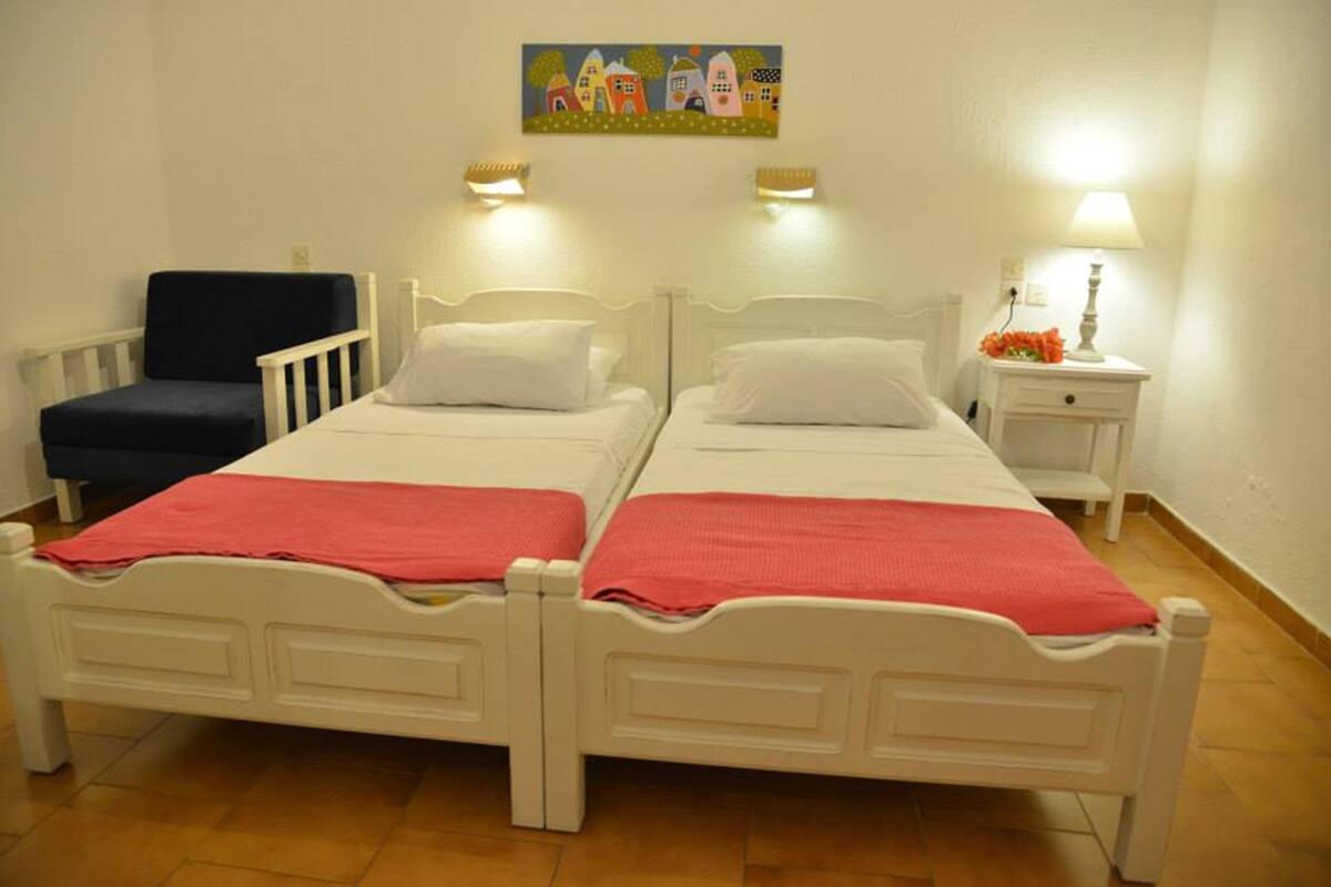 Una habitación con dos camas blancas, una silla azul, una mesita con una lámpara y un cuadro en la pared.