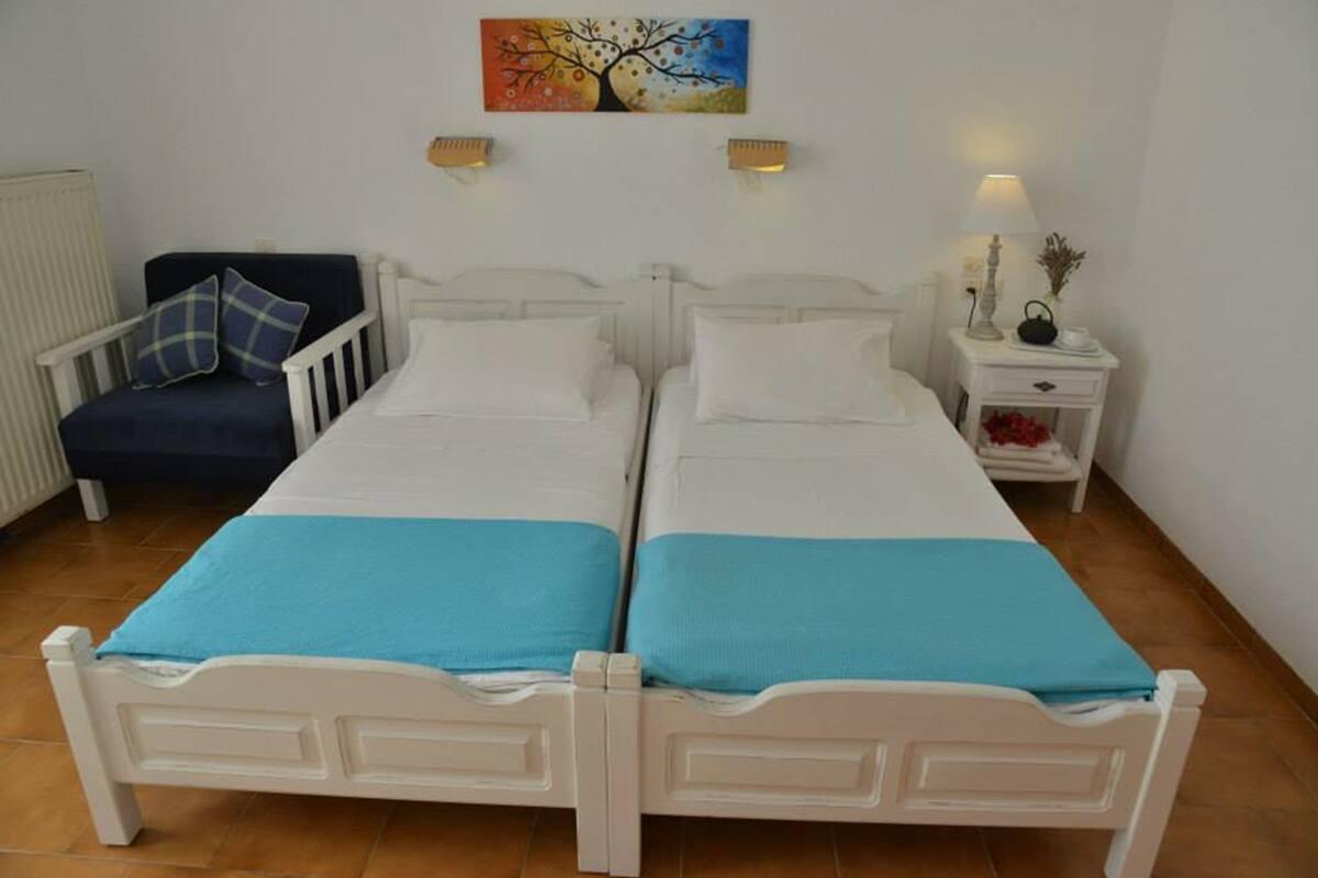 Una habitación con dos camas individuales, una silla, una mesita de noche con lámpara y un cuadro en la pared.