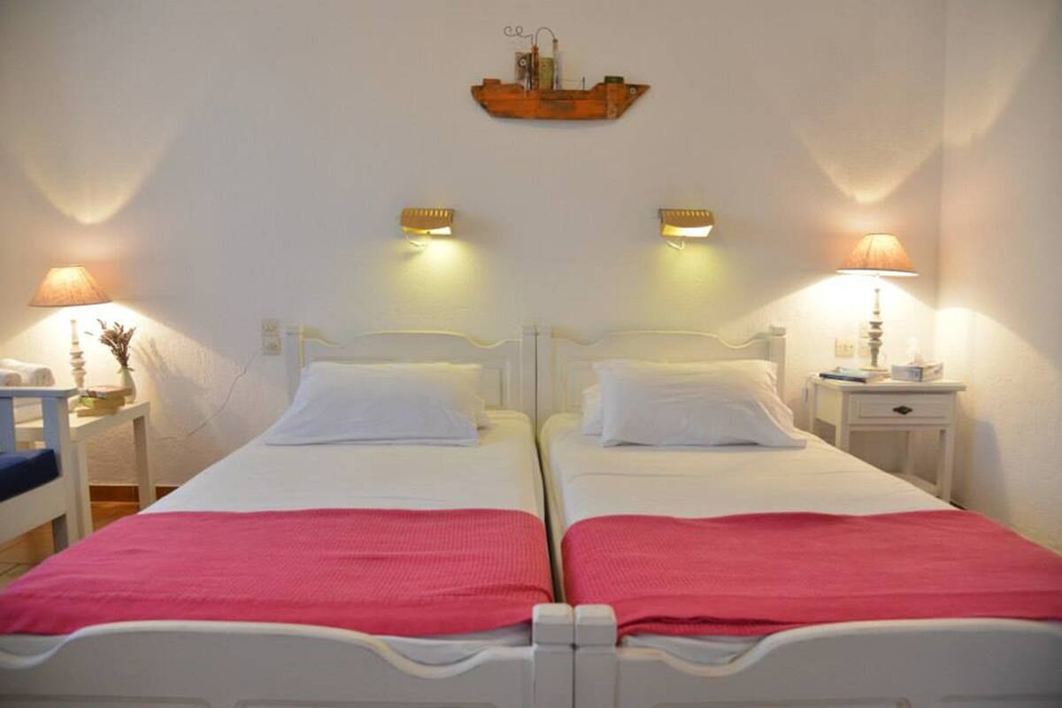 Habitación de hotel con dos camas, cada una con cabecero blanco y manta rosa. Mesitas de noche con lámparas y un pequeño objeto decorativo en cada una.