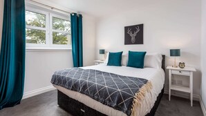 A bedroom with a bed, teal curtains, a wall-mounted clock, and a framed deer head artwork. - Elms Way - Donnini Apartments (Ayr)