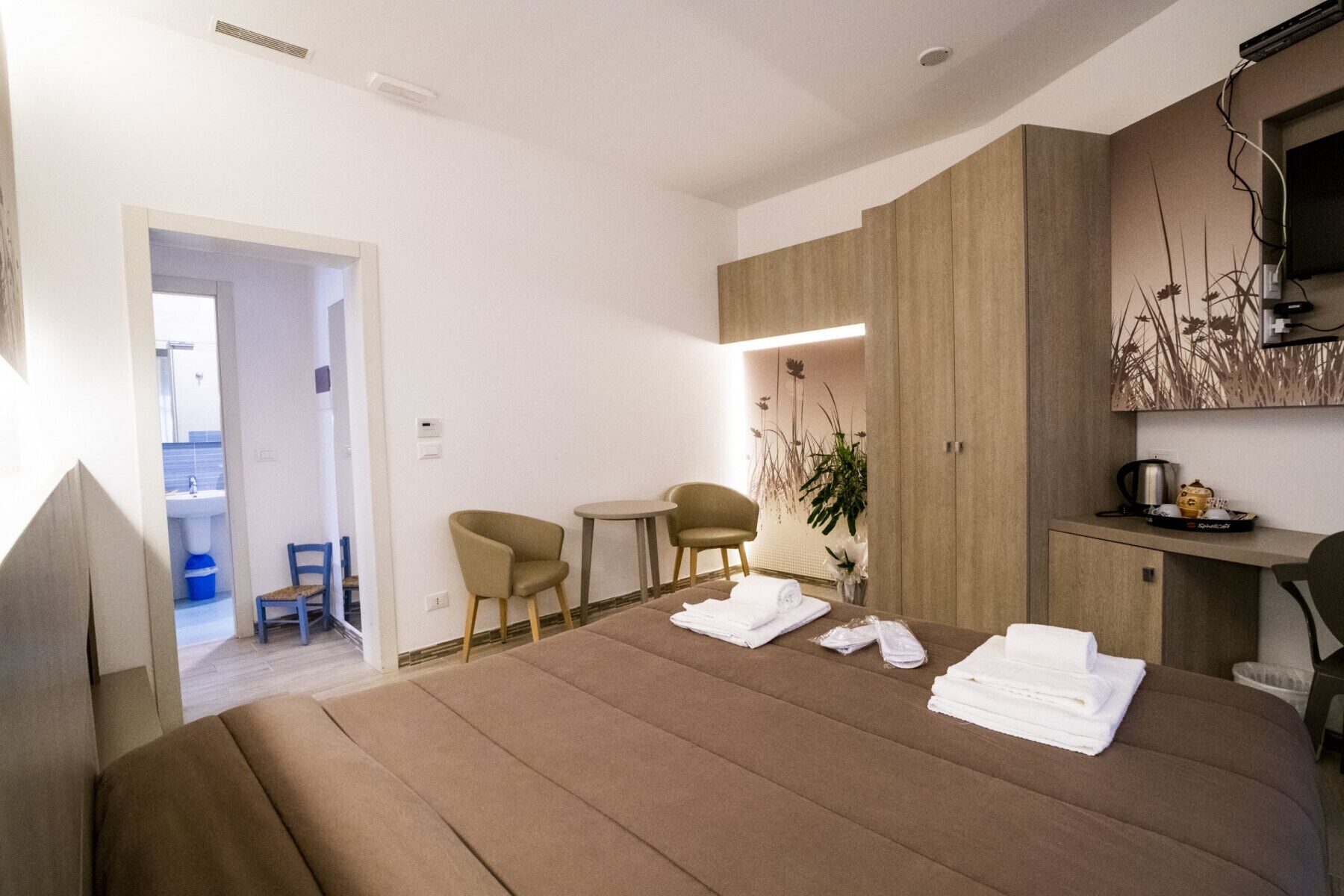 Una habitación de hotel moderna con una cama grande, un área de estar con dos sillones, una mesita y una kitchenette.