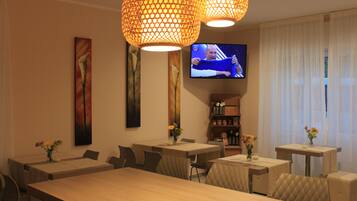 Comedor de hotel con varias mesas y sillas, un televisor montado en la pared y lámparas decorativas.