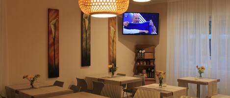 Comedor de hotel con varias mesas y sillas, un televisor montado en la pared y lámparas decorativas.