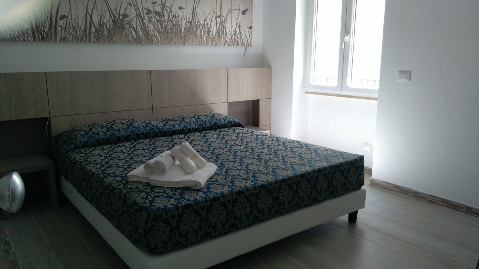 Una cama bien hecha con una colcha estampada y toallas enrolladas, una mesita de noche y una ventana con luz natural.