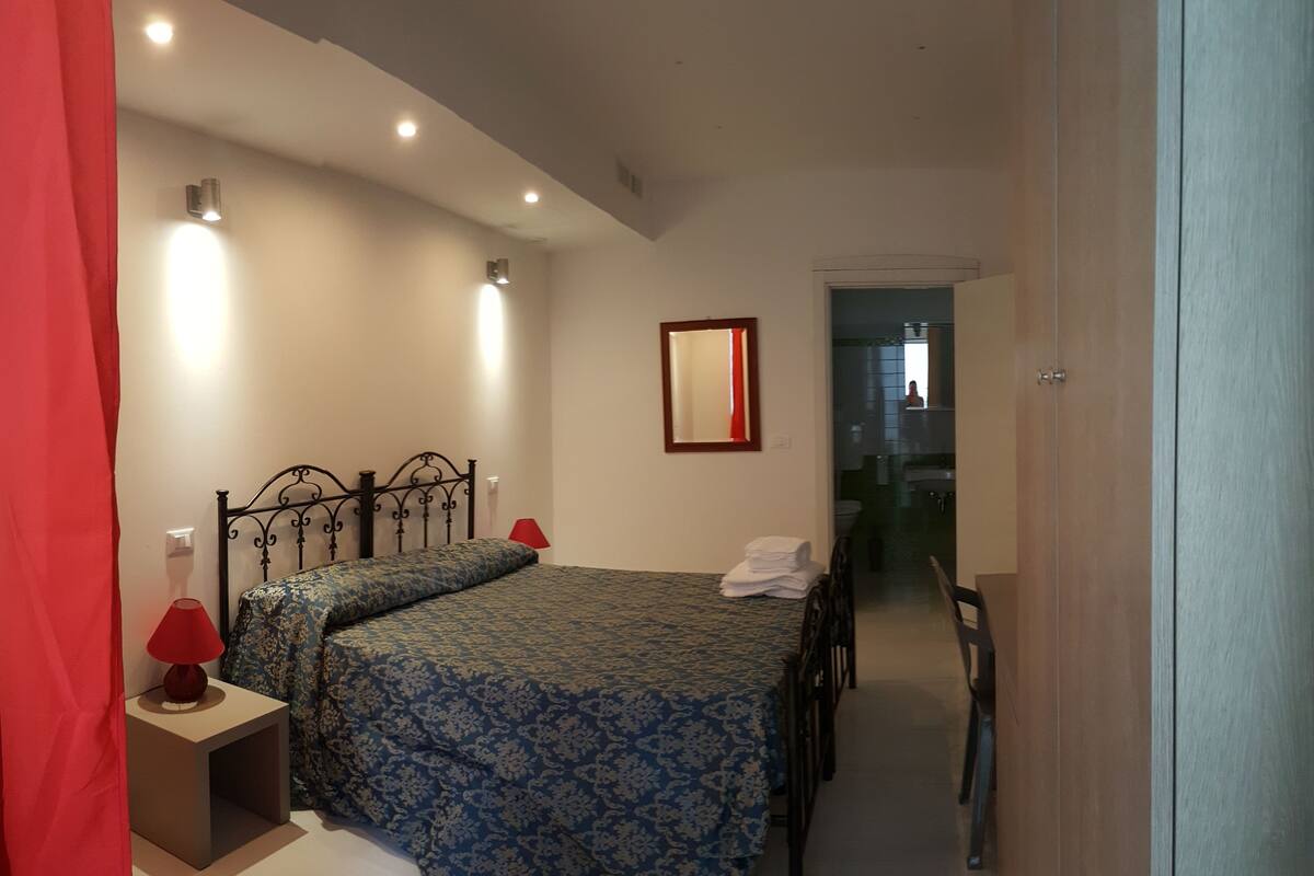 A bedroom with two beds, a bedside table with a lamp, a mirror on the wall, and a bathroom visible through an open door.