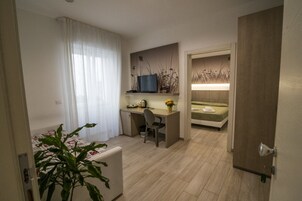 A hotel room with a wooden floor, a desk with a chair, a bed, a television on the wall, and a potted plant.