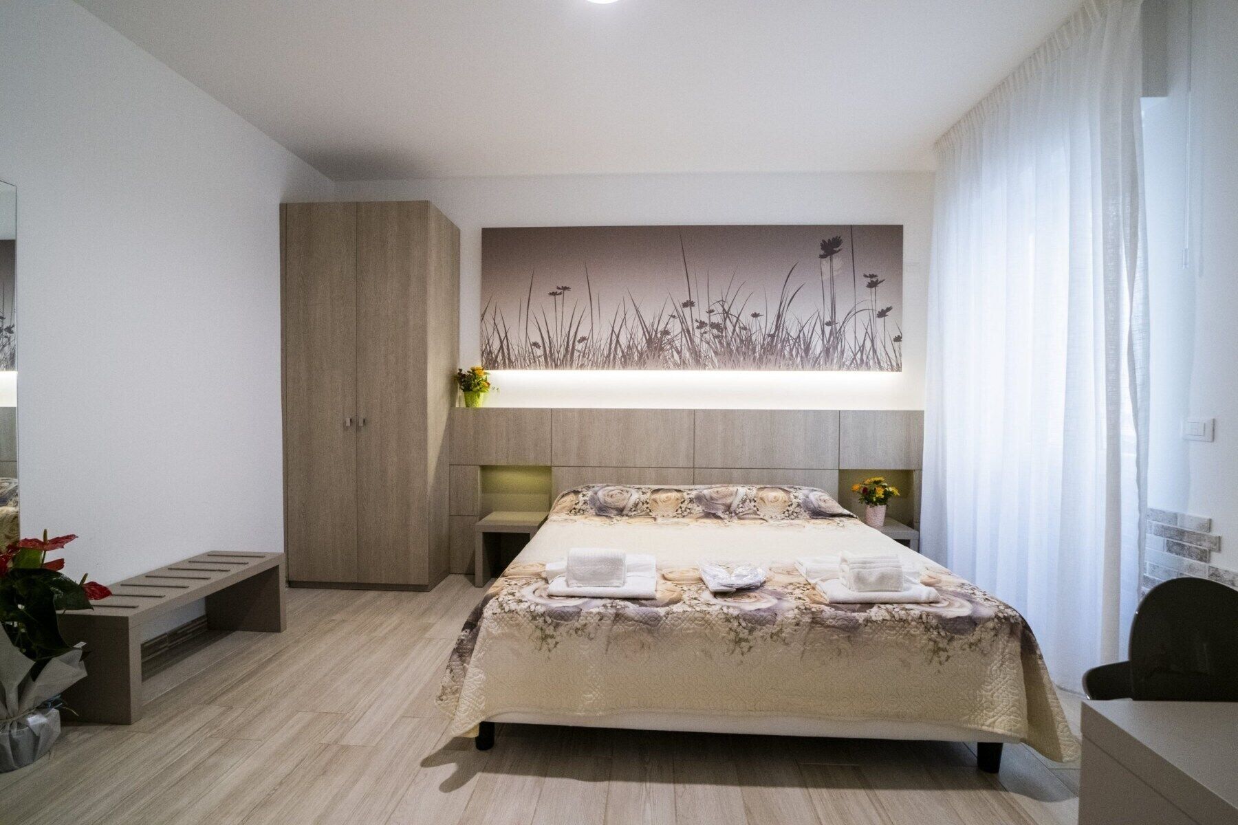 Una camera d'albergo moderna con un letto grande, un armadio in legno, un comodino con una lampada e un'opera d'arte appesa al muro.