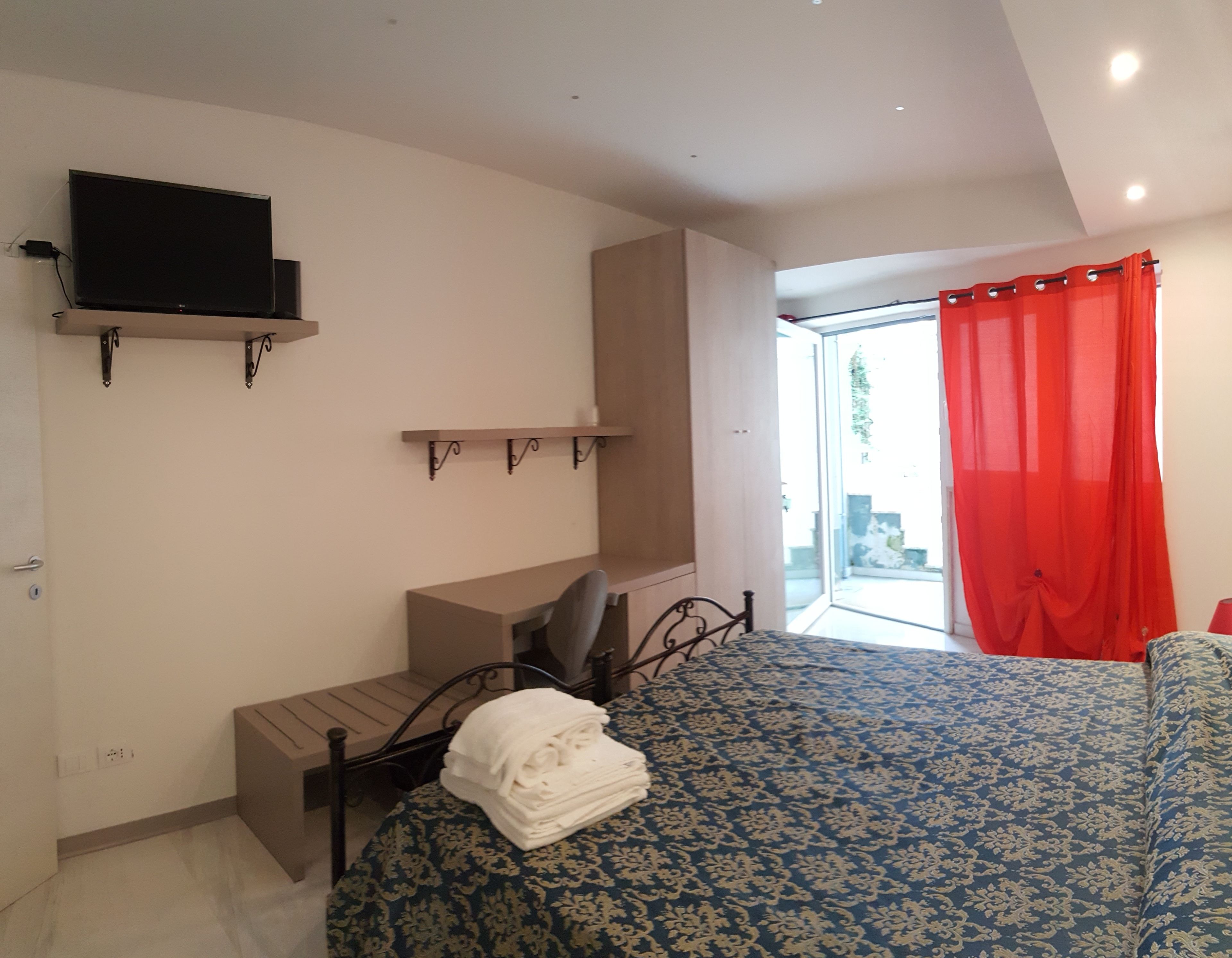 A bedroom with a bed, a desk, a chair, a TV mounted on the wall, and a window with red curtains.