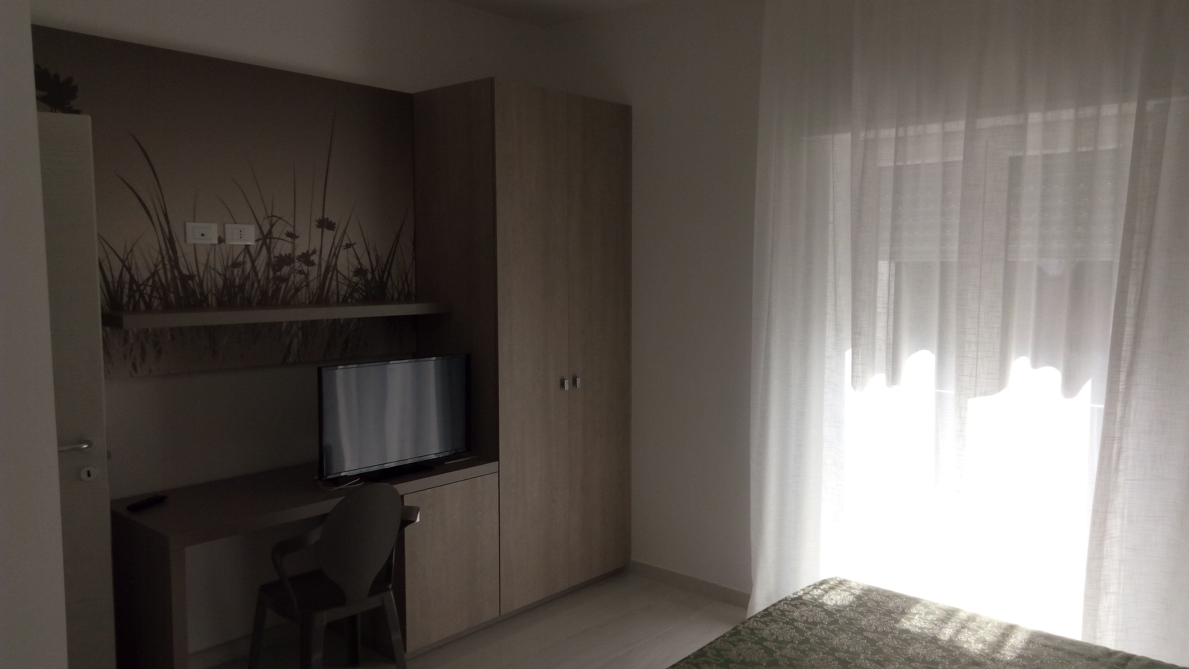 Ein Schlafzimmer mit einem Bett, einem Schreibtisch mit Fernseher, einem Stuhl und einem Fenster mit leichtem Vorhang.