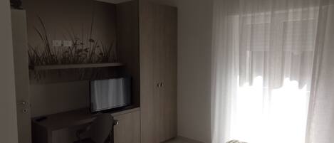 Ein Schlafzimmer mit einem Bett, einem Schreibtisch mit Fernseher, einem Stuhl und einem Fenster mit leichtem Vorhang.