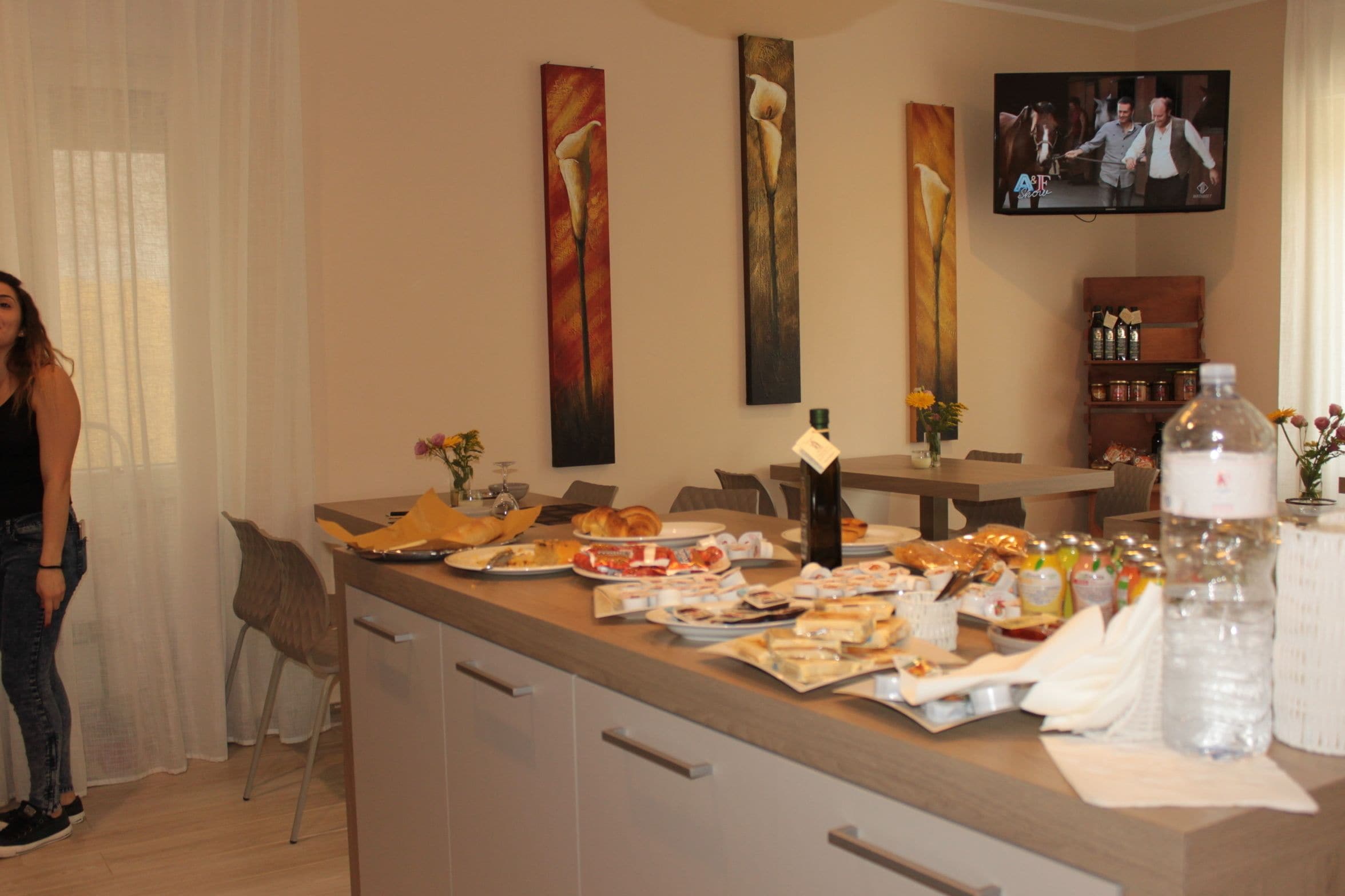 A hotel room with a dining area featuring a buffet setup with various dishes and drinks. There are paintings on the wall, a television, and a minibar.