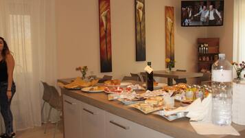 A hotel room with a dining area featuring a buffet setup with various dishes and drinks. There are paintings on the wall, a television, and a minibar.