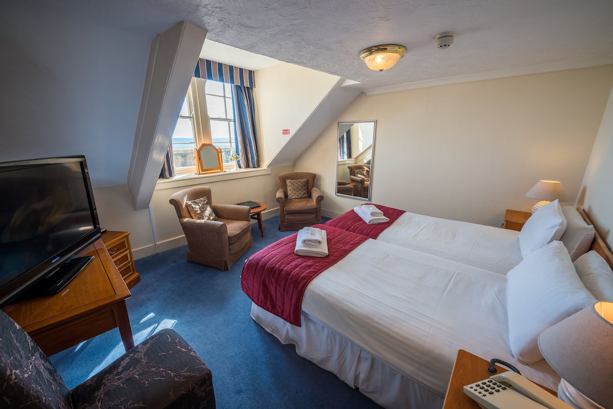 A hotel room with a large bed, a desk with a TV, a chair, a small table, and a window with curtains.