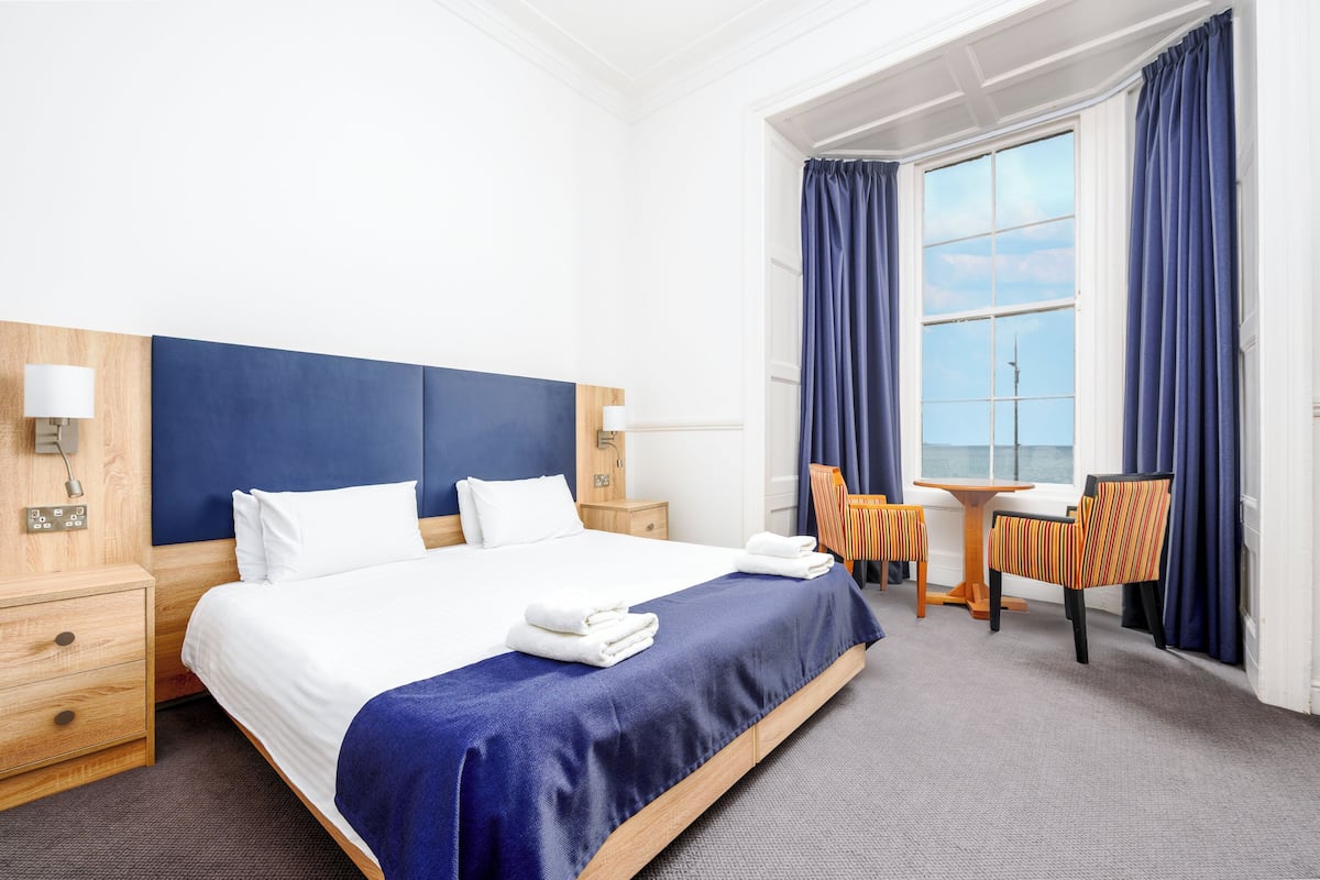 A hotel room with a large bed, a wooden nightstand, a desk with a chair, and a view of the sea through a window.