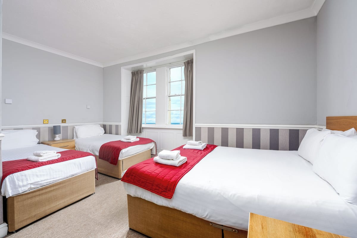 A hotel room with three beds, a window with curtains, and a view of the sky.