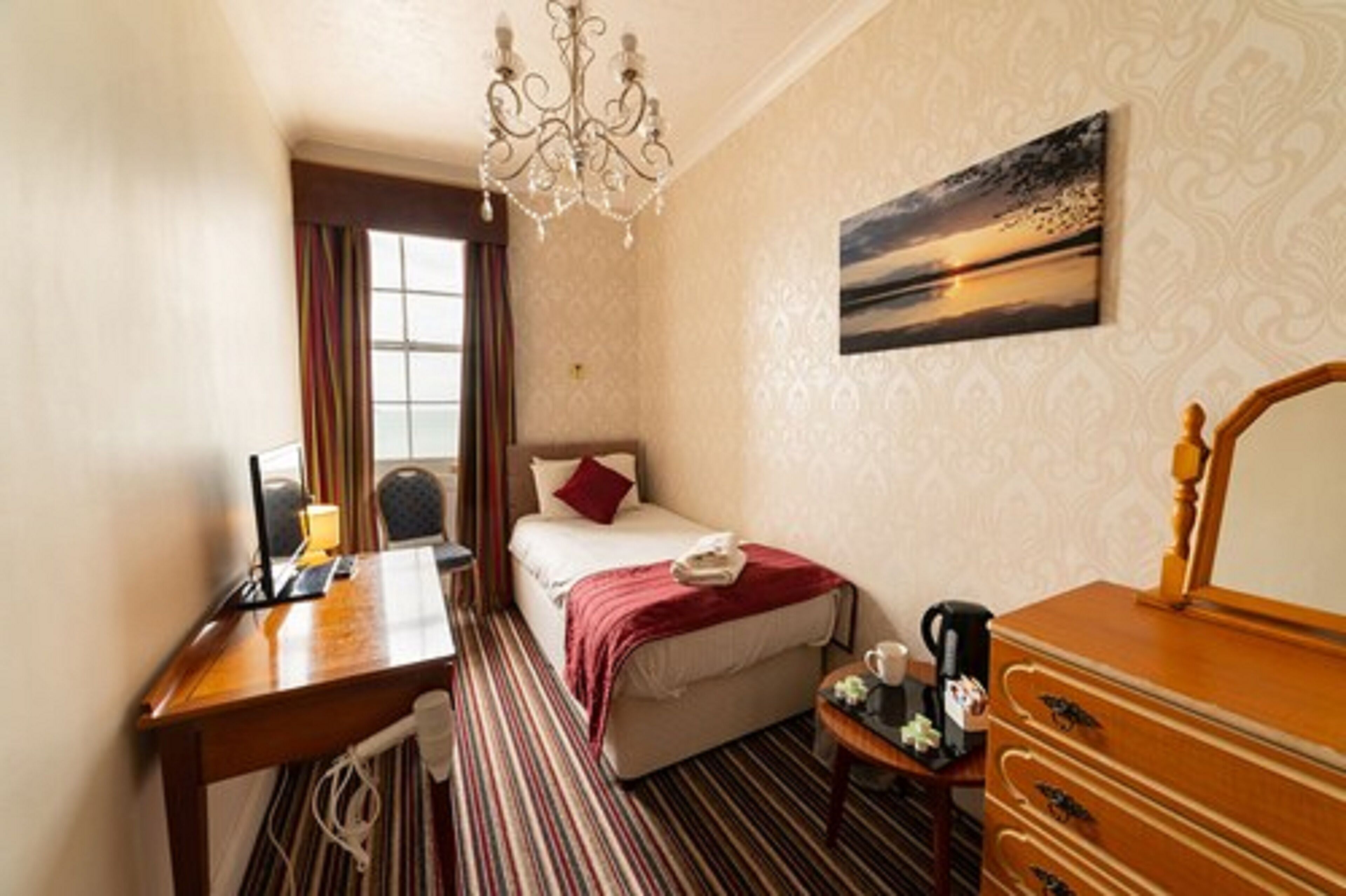 Habitación de hotel con cama, escritorio, cómoda, candelabro, pintura y ventana con cortinas.