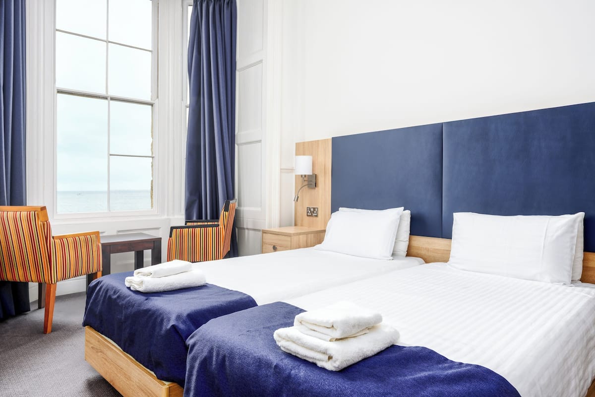 A hotel room with two beds, a wooden headboard, a nightstand, a lamp, a chair, and a view of the sea.