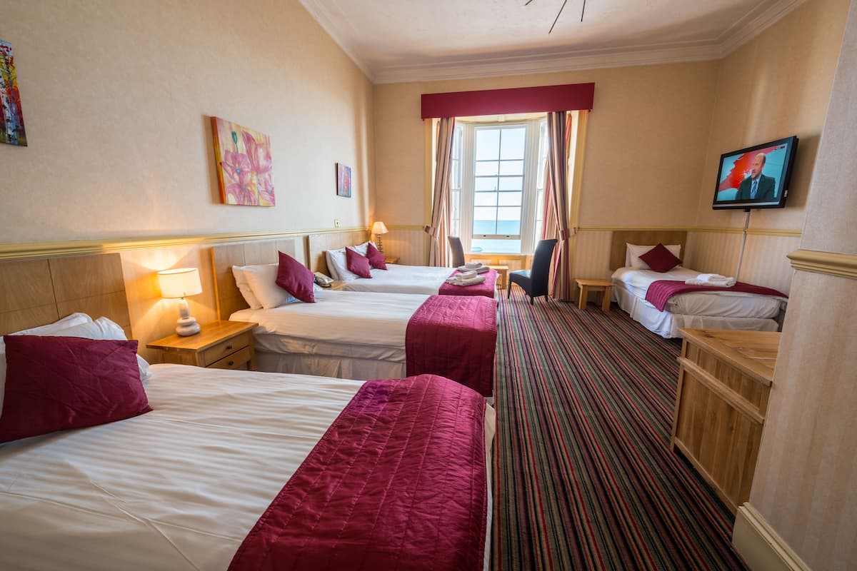 A hotel room with three beds, a television, a window with curtains, and artwork on the walls.