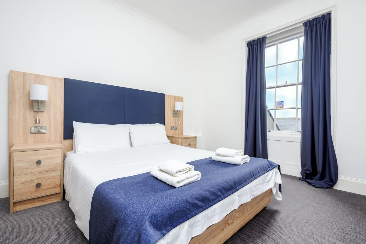A neatly made bed with a blue bedspread, white pillows, and two folded towels on the side. There is a wooden nightstand with a lamp on each side of the bed.