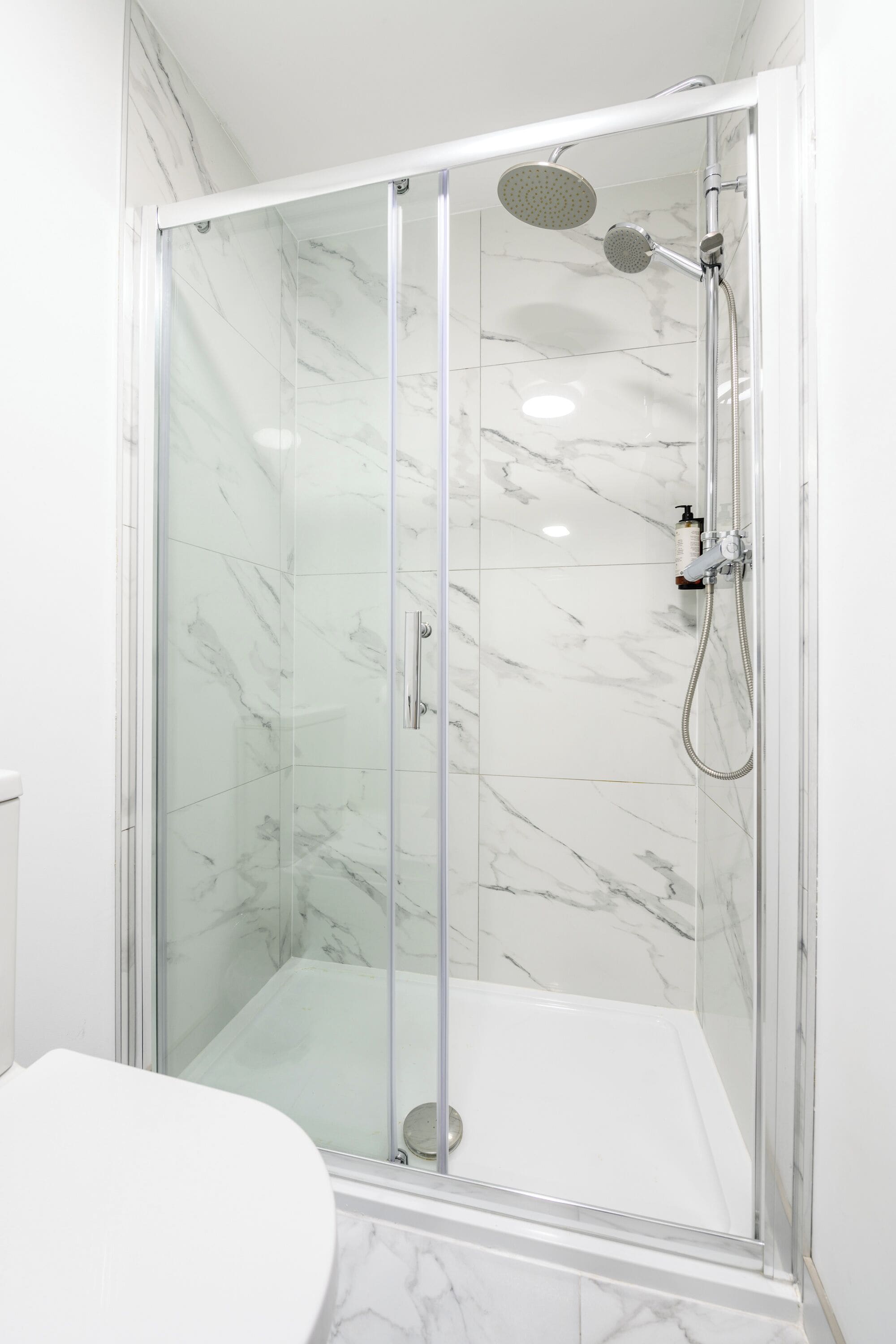 Een douchekabinet met glazen wanden, een witte marmeren wand en vloer, en een handdouche met een spuitmond.