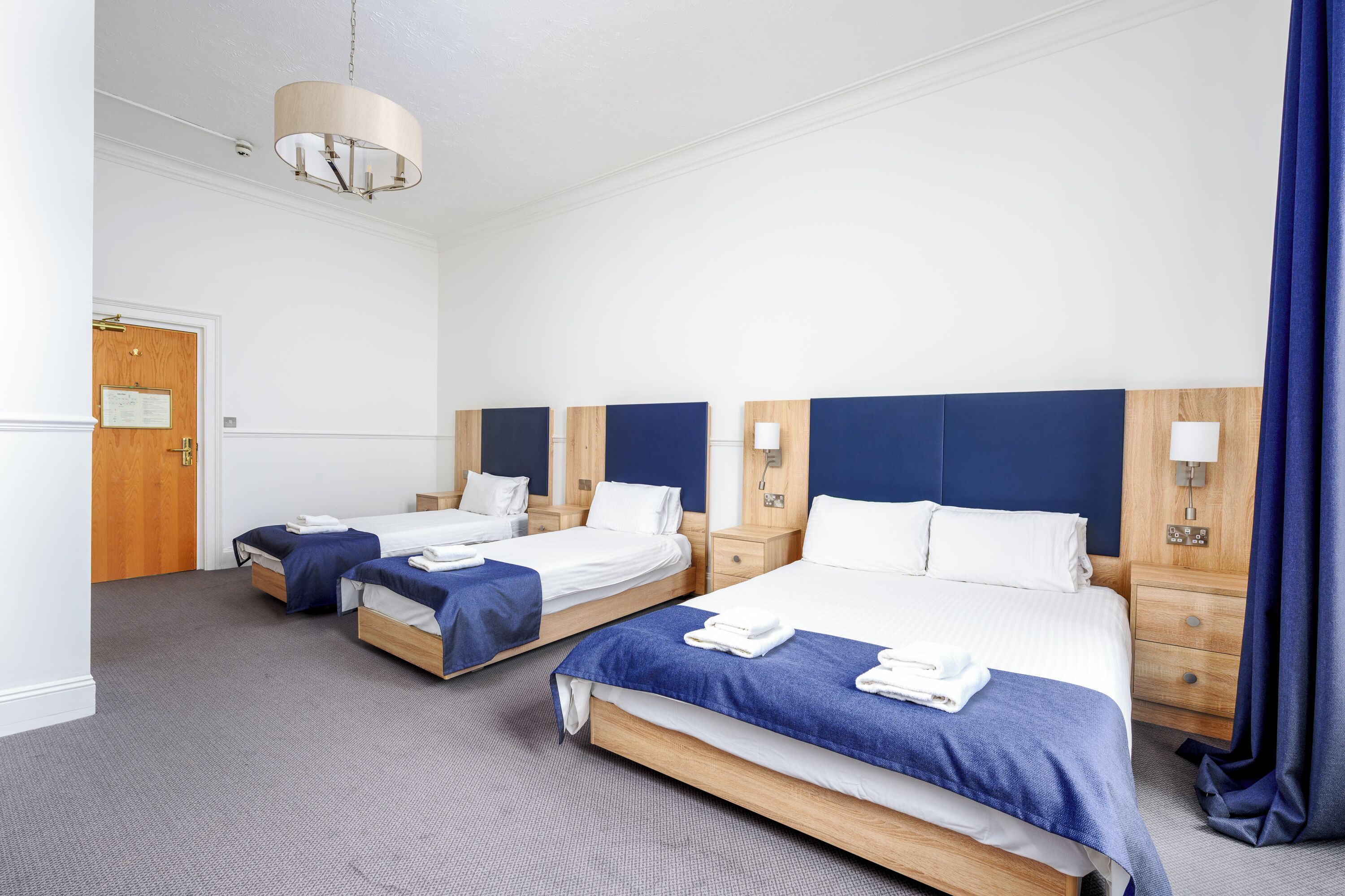 A hotel room with three beds, a wooden door, a blue curtain, and a hanging light fixture.