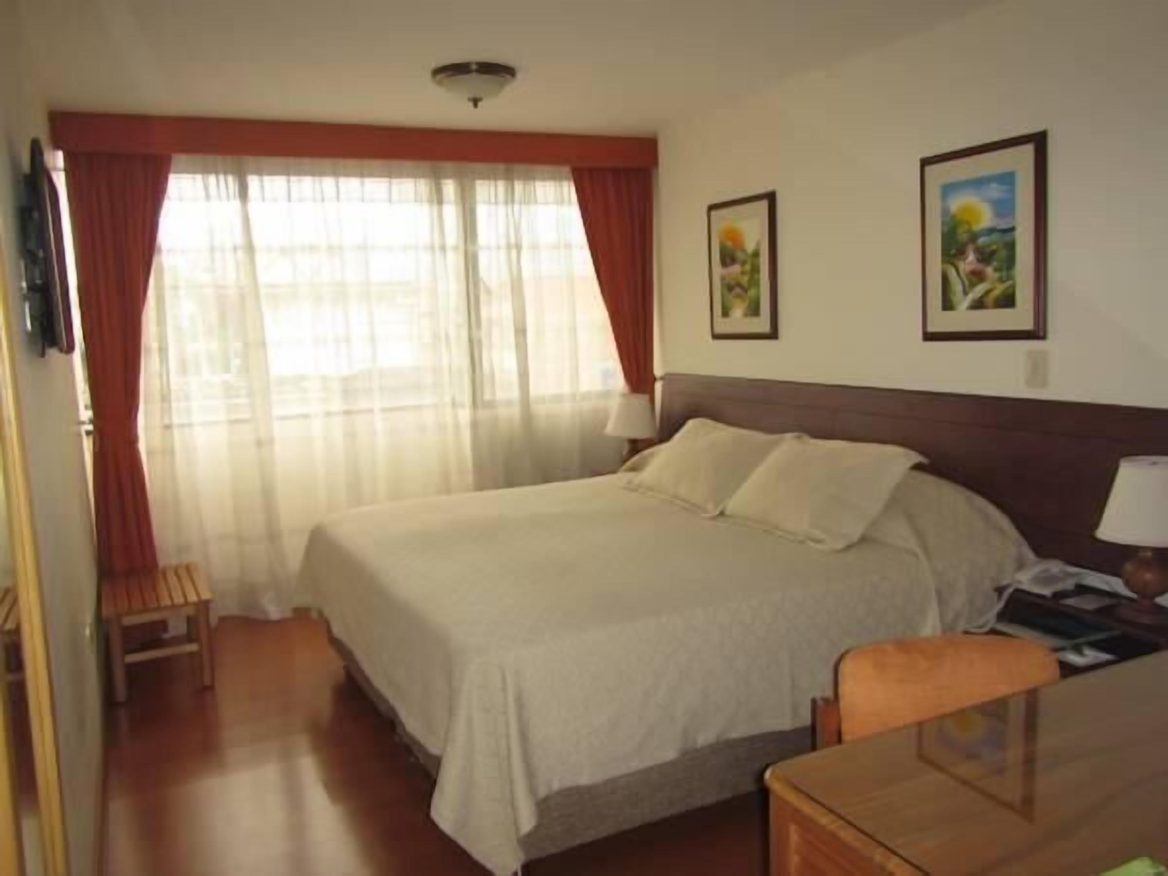 A hotel room with a large bed, a wooden chair, a desk, and two framed pictures on the wall.