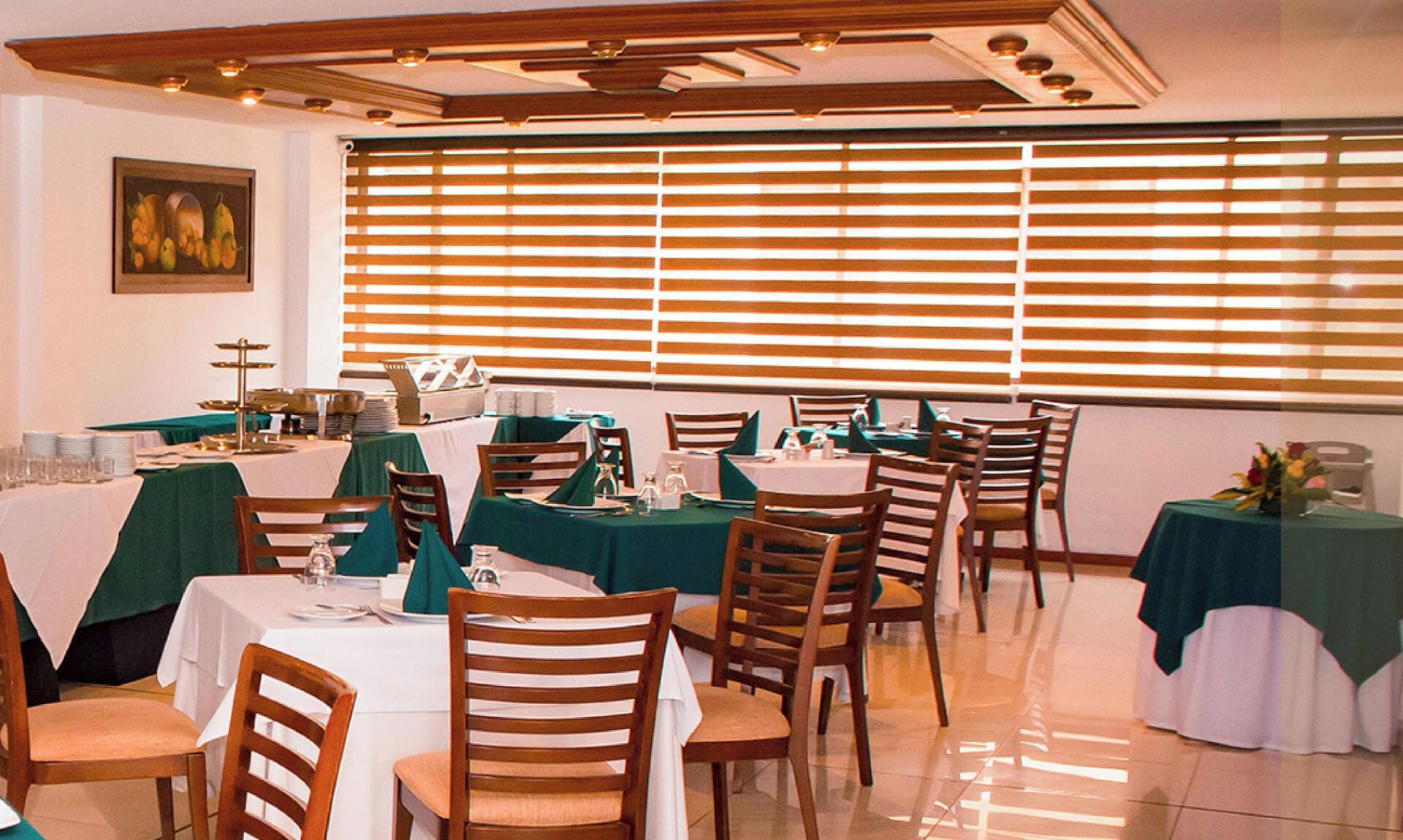 Un comedor con mesas dispuestas para el servicio, sillas de madera y un ventanal con persianas horizontales.