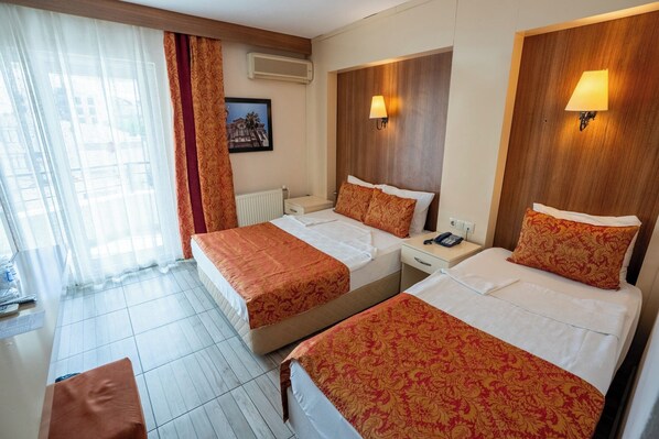 A hotel room with two beds, a window with sheer curtains, a radiator, a bedside table with a phone, and a wall-mounted lamp. - Buyuk Berk Otel (Ayvalik)