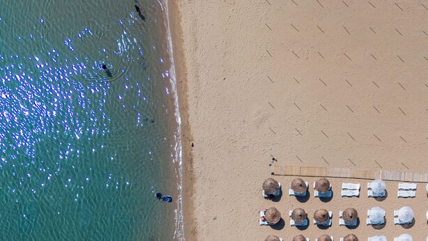 Vista aérea de uma praia com fileiras de espreguiçadeiras e guarda-sóis, uma torre de salva-vidas e algumas pessoas na água.