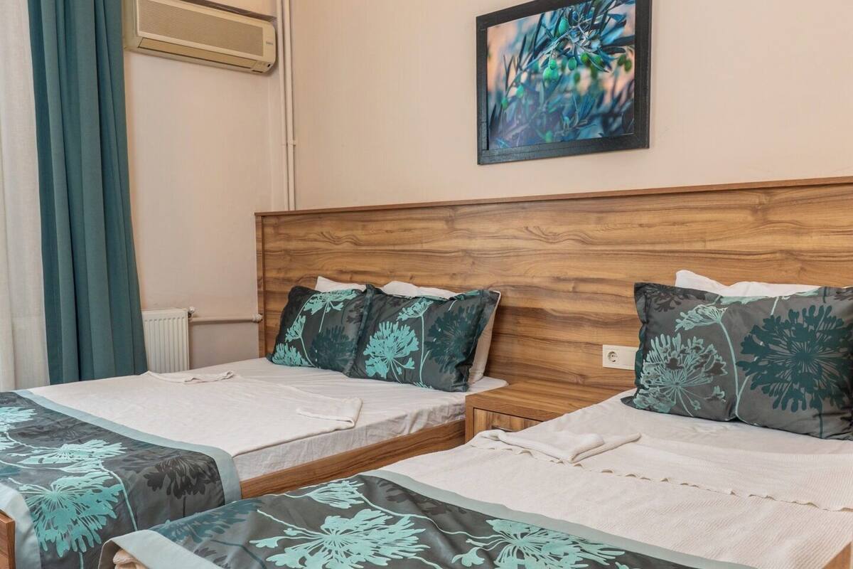 A hotel room with two wooden beds, a large window with curtains, a wall-mounted air conditioner, and a framed picture on the wall.