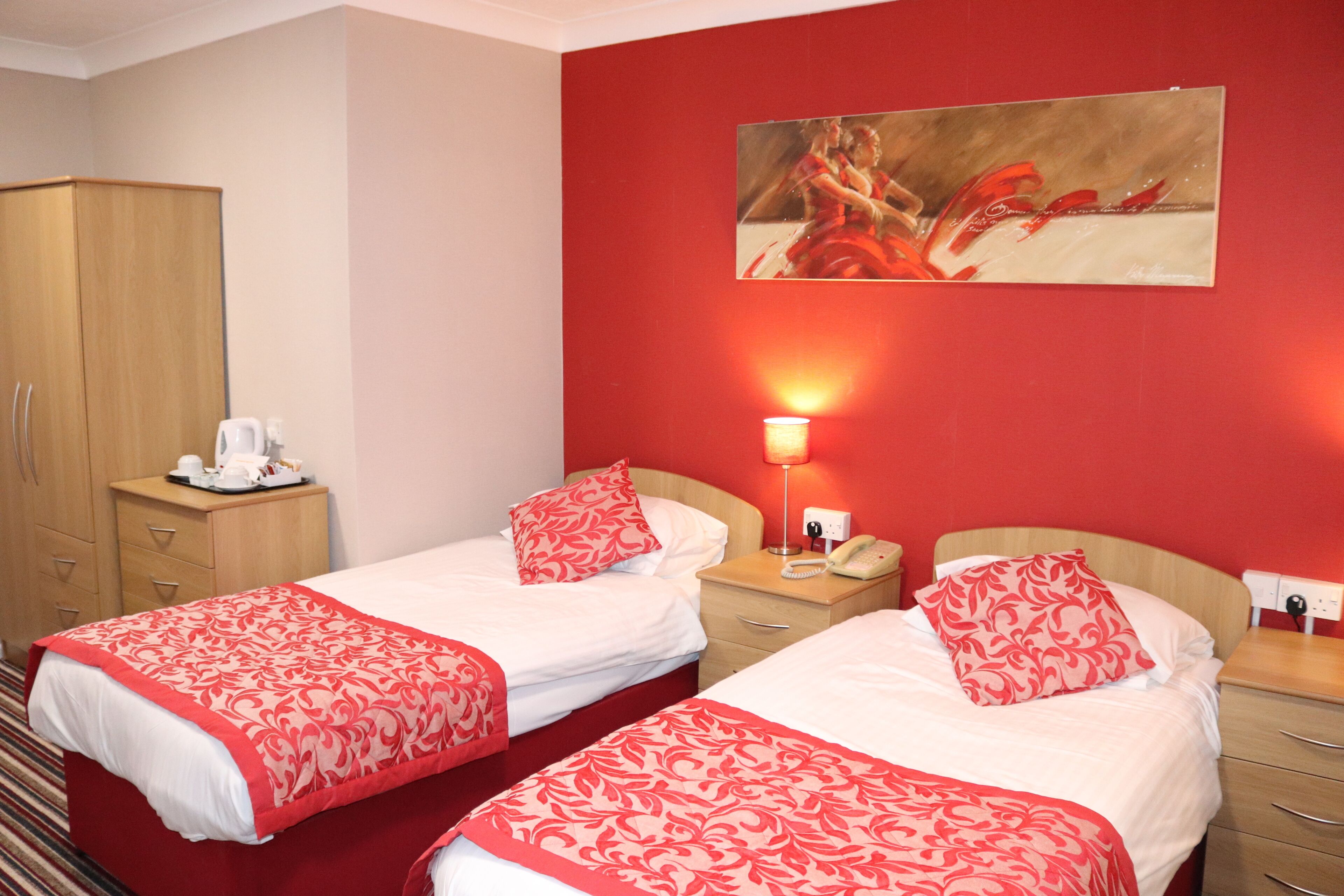 A hotel room with two beds, a wooden wardrobe, a painting on the wall, and a nightstand with a lamp and phone.