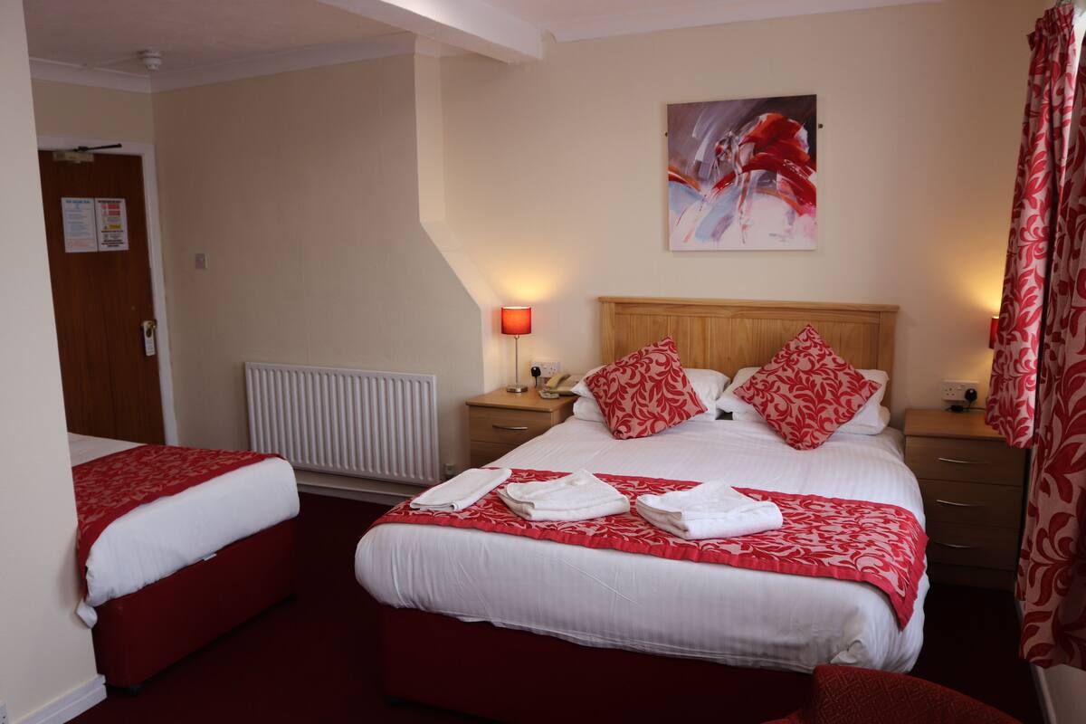 A hotel room with two beds, a wooden headboard, a red and white patterned bedspread, a red and white pillow, a nightstand with a lamp, and a painting on the wall.