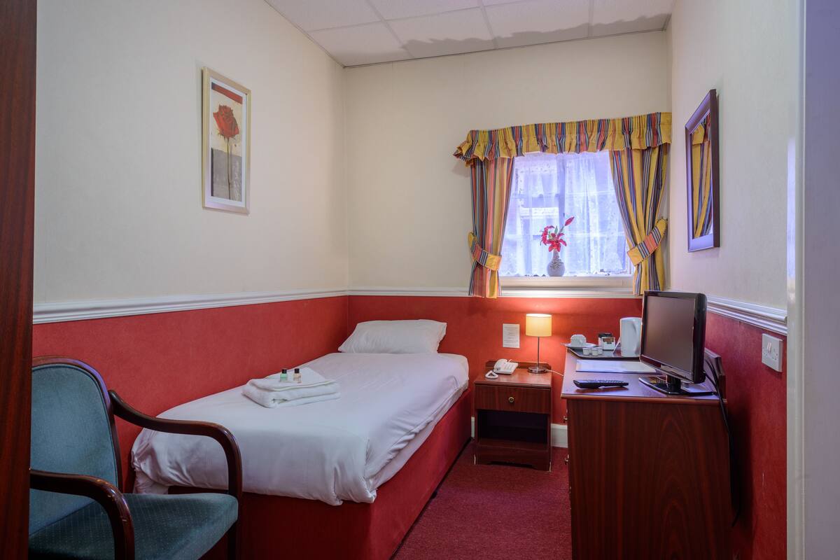 A hotel room with a single bed, a desk with a computer and phone, a chair, and a window with curtains.