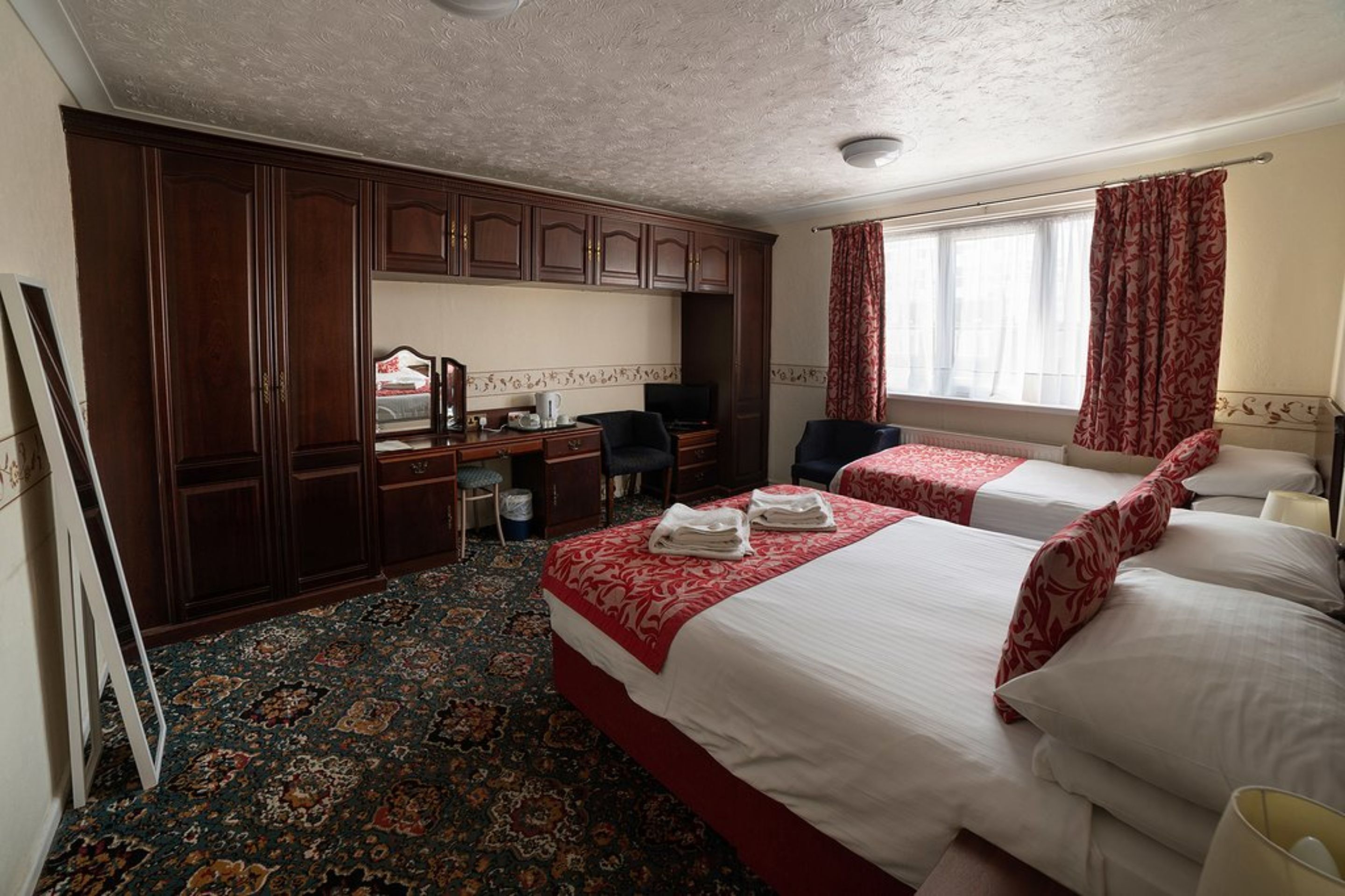A hotel room with two beds, a desk with a chair, a wardrobe, and a large window with curtains.