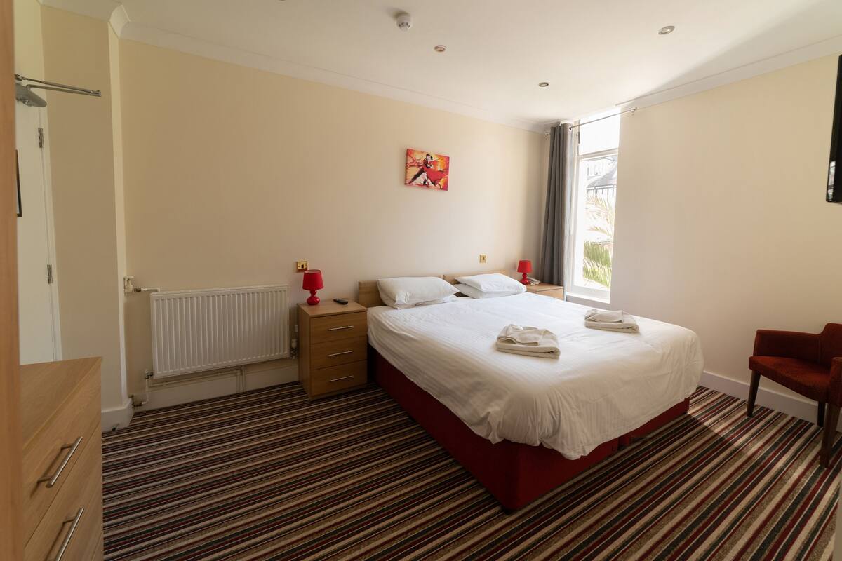 A hotel room with a bed, a red chair, a radiator, a wooden dresser, and a window with curtains.