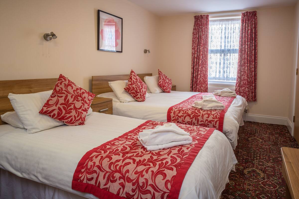 A hotel room with two beds, each with red and white bedding, a wooden headboard, and a red patterned carpet.