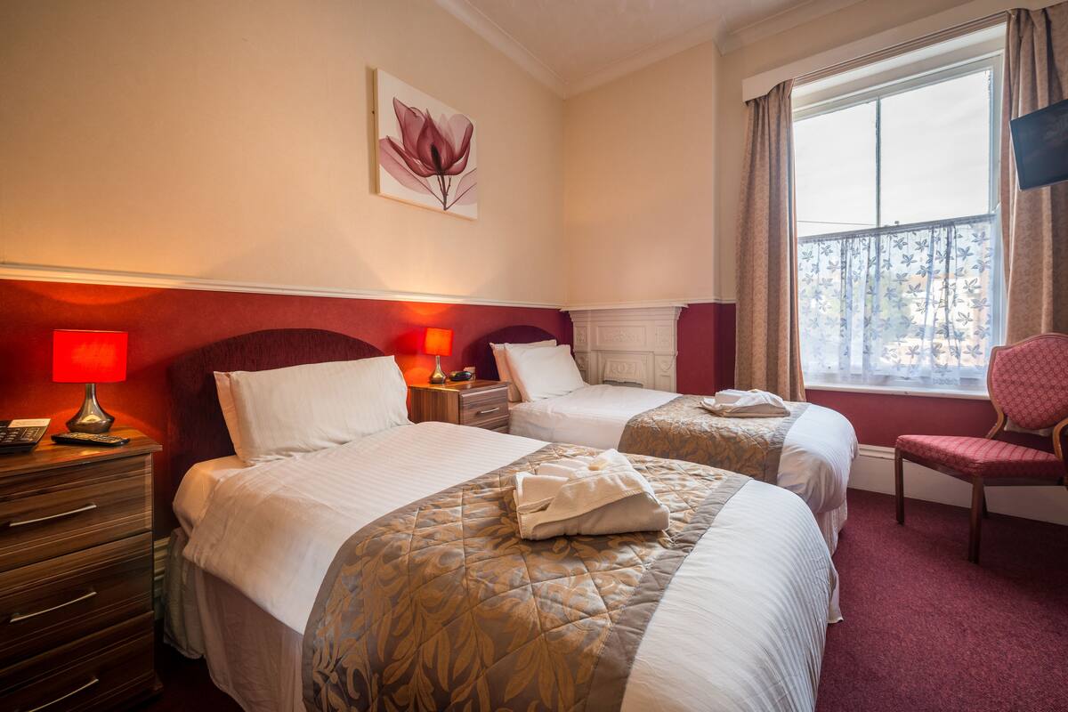 A hotel room with two beds, a red wall, a painting, a window with curtains, a chair, and a nightstand with a lamp.