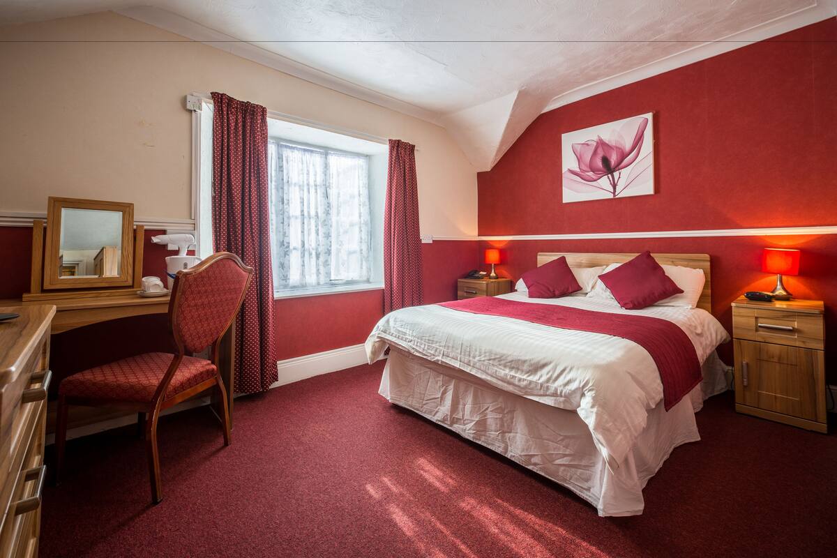 A bedroom with a bed, a desk, a chair, a nightstand, and a window with curtains.
