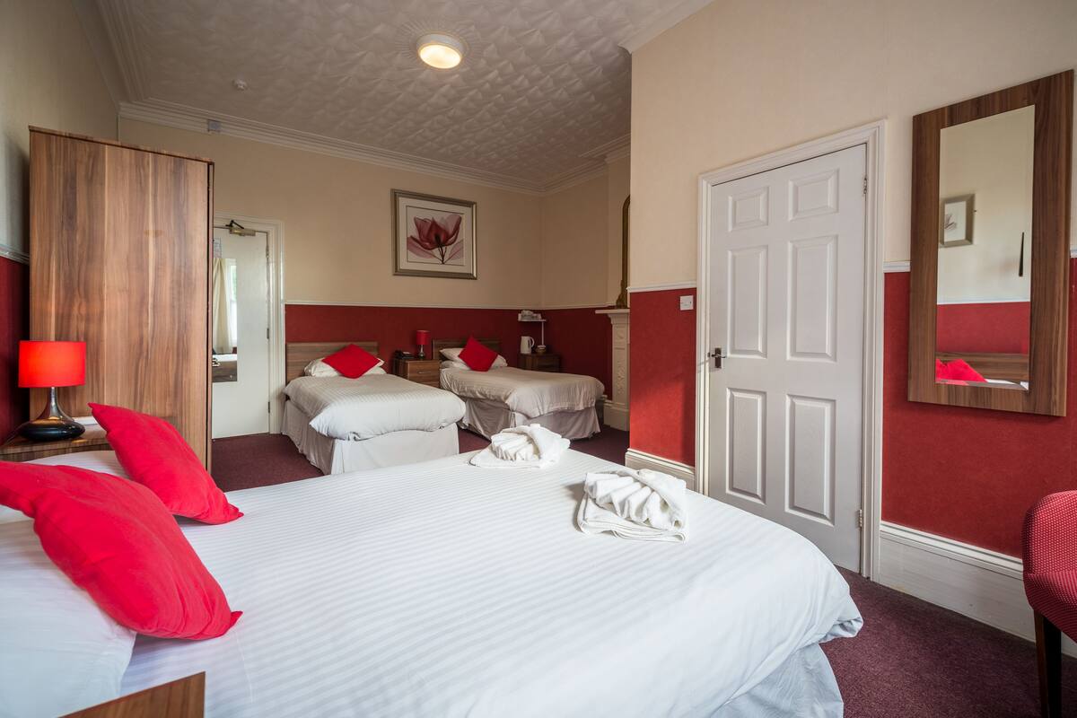 A hotel room with a large bed, two smaller beds, red pillows, a red lamp, a mirror, and a framed picture on the wall.