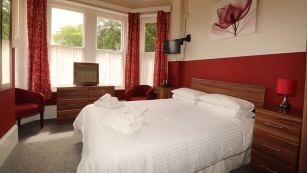 Quarto de hotel com uma cama grande, uma mesa com televisão, uma cadeira vermelha e um abajur vermelho.