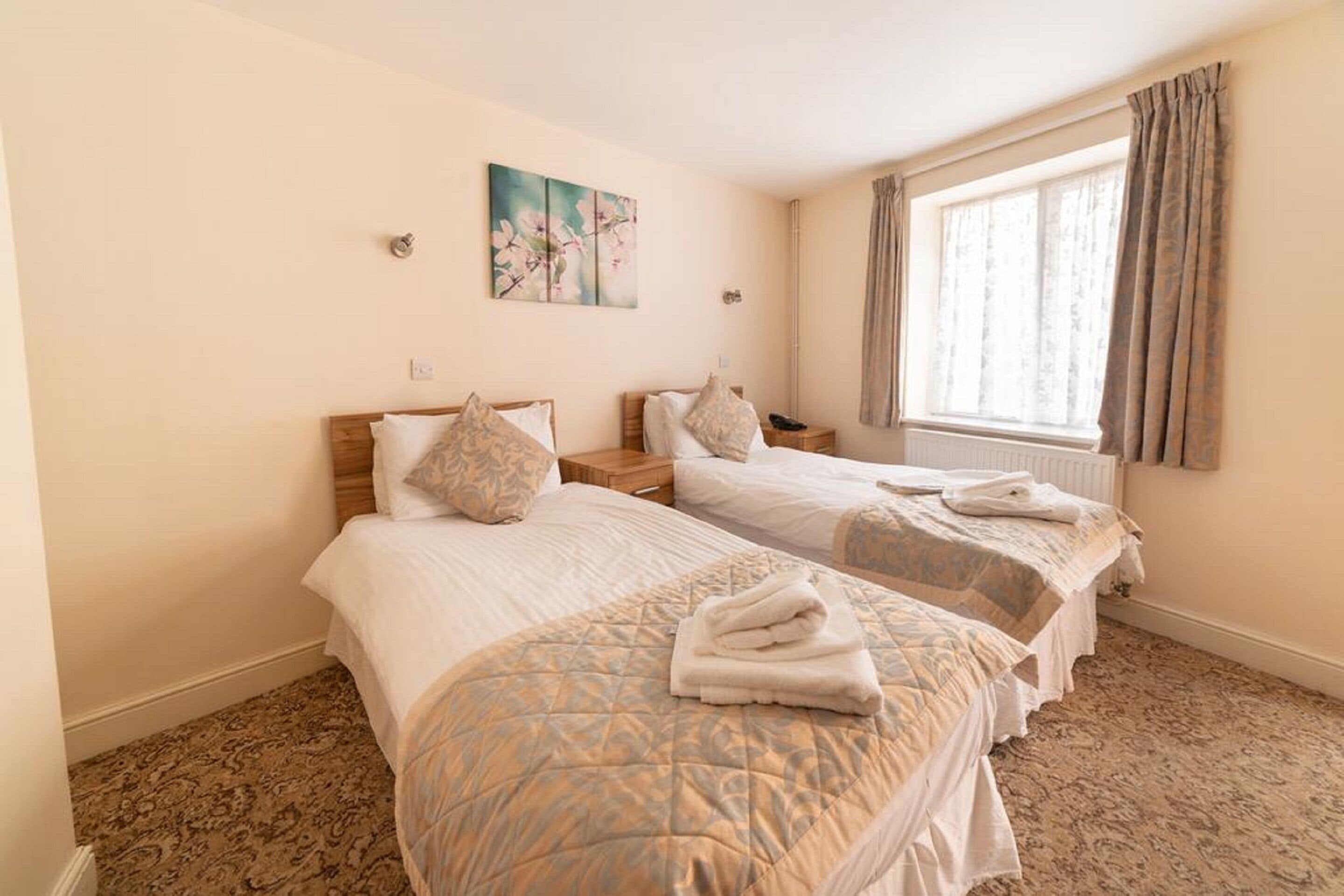A hotel room with two beds, a painting on the wall, a window with curtains, and a radiator.