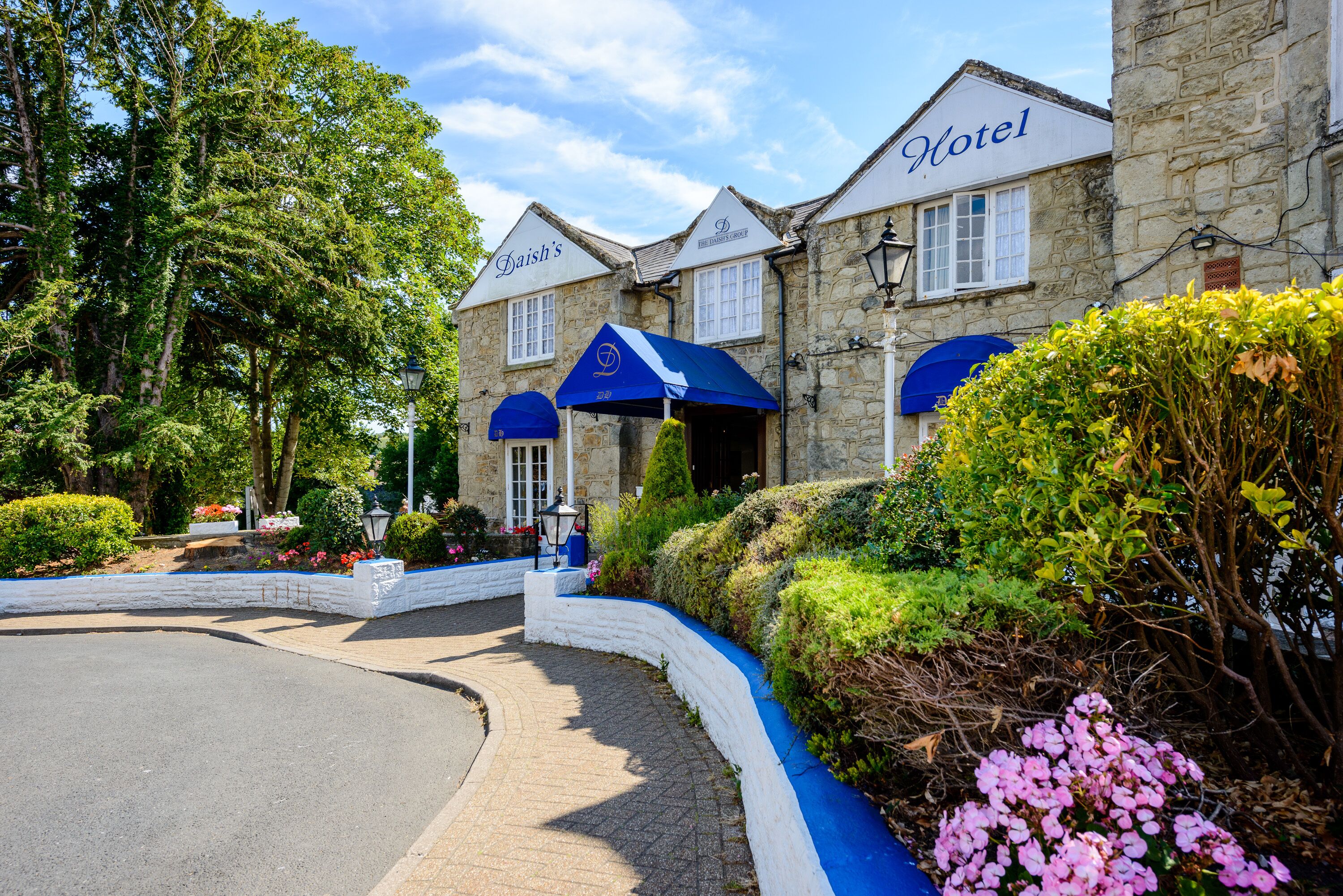 Ein Hotel mit Stein-Fassade, zwei blauen Markisen und einem angelegten Garten.