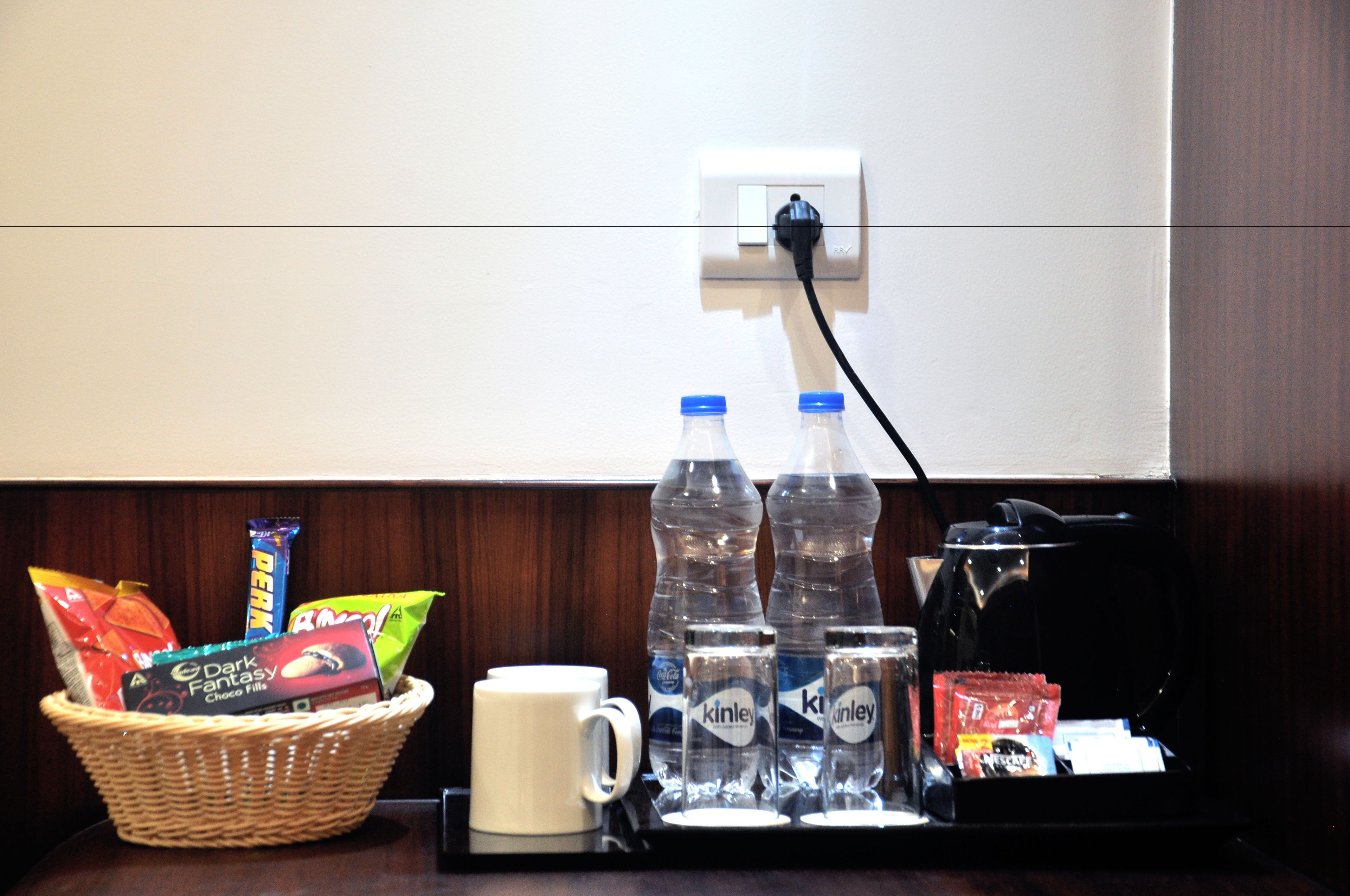 A hotel room with a wicker basket containing snacks and bottled water, a white mug, and a coffee maker on the counter.