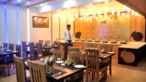 A restaurant with wooden tables and chairs, a buffet counter, and a waiter. - Hotel SGT Plaza (Varanasi)