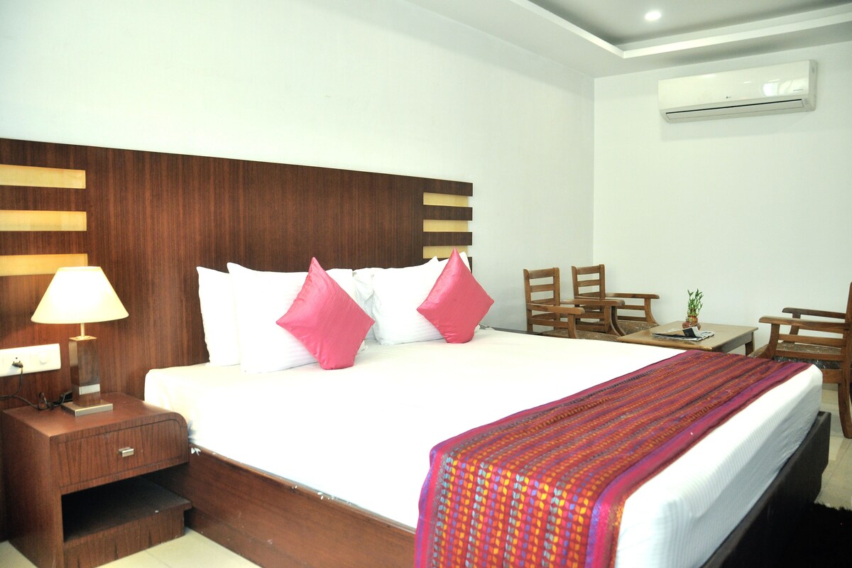 A hotel room with a large bed, a wooden nightstand with a lamp, a small table with a plant, and a chair.