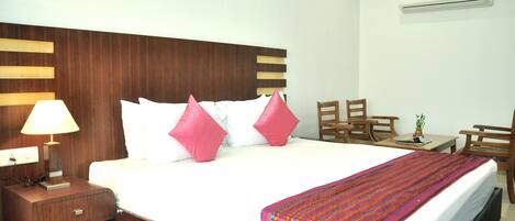 A hotel room with a large bed, a wooden nightstand with a lamp, a small table with a plant, and a chair.