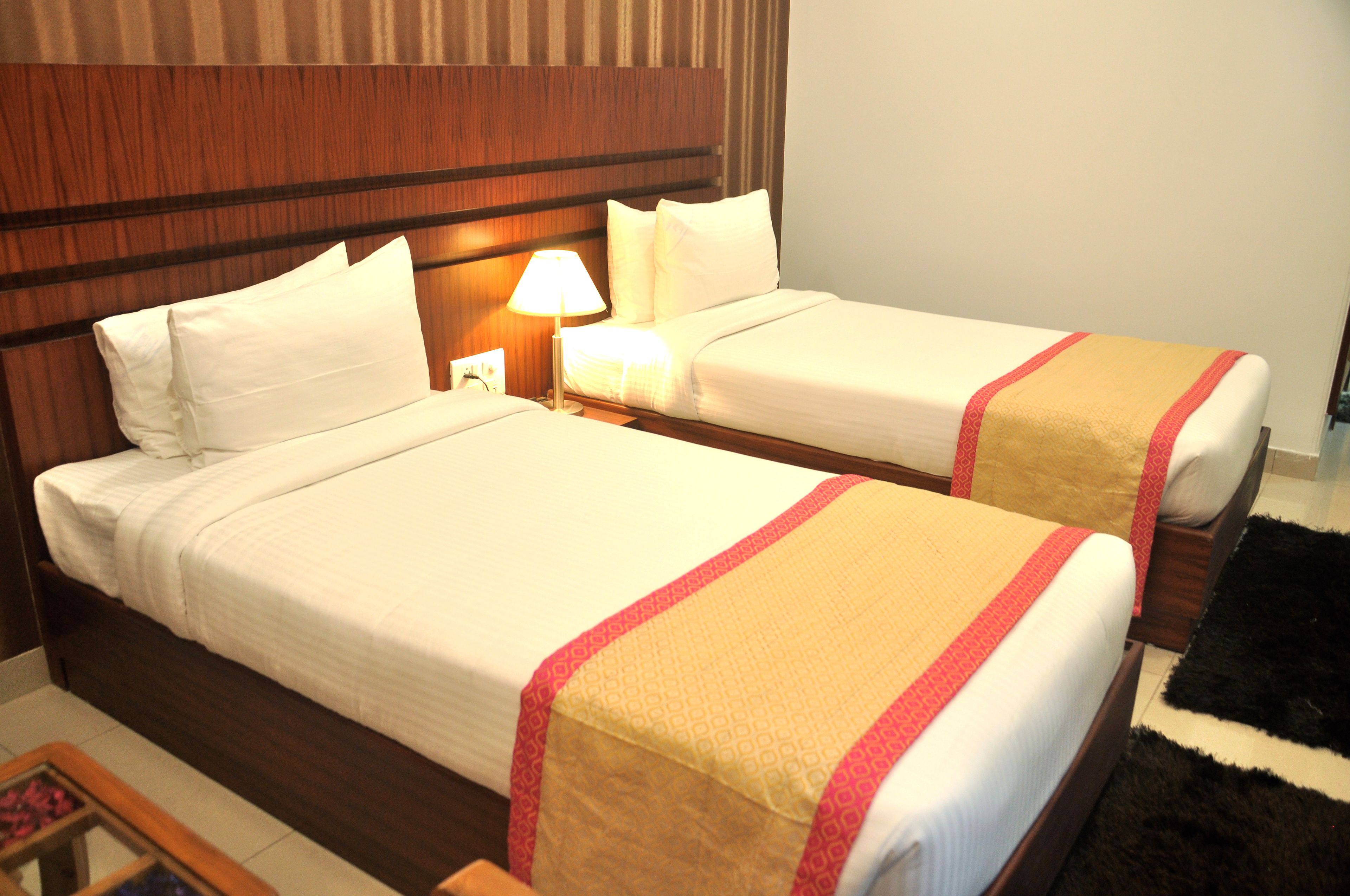 Ein Hotelzimmer mit zwei Betten, jeweils mit weißer Bettwäsche und einer roten und beigen Tagesdecke. Dazwischen befindet sich ein Nachttisch mit einer Lampe.